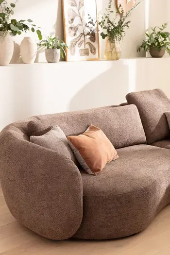 Linke abgerundete Armlehne und teile der Sitzfläche eines braunen Sofas, auf dem Kissen liegen in beige und orange und viele Pflanzen dahinter.